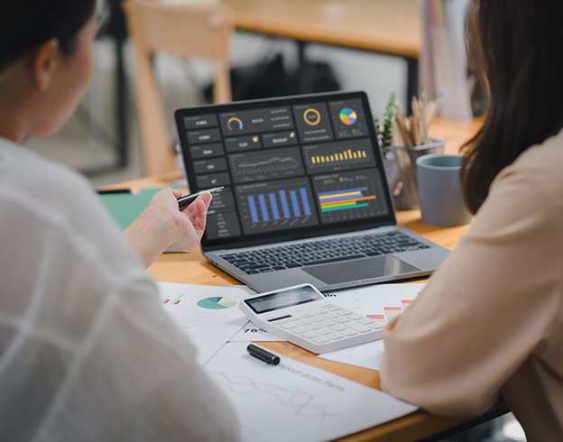 Two people analyzing data on a laptop with charts, graphs, documents, and a calculator on the table.